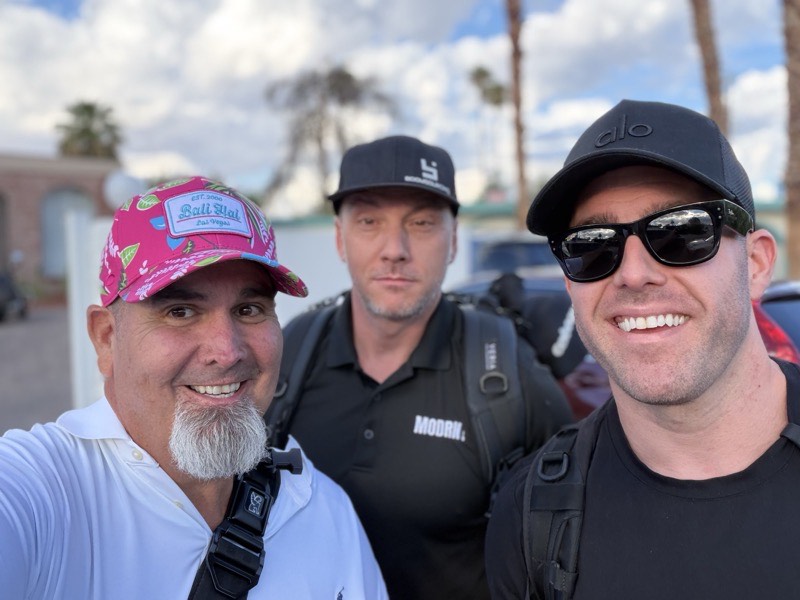 Three people outdoors in daylight with palm trees behind them.