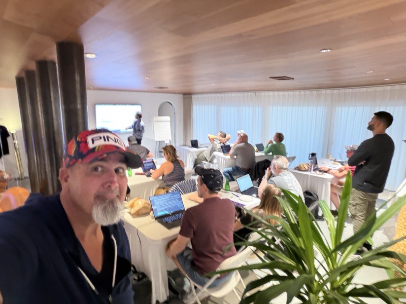 People working on laptops at tables in a daylit room with a large plant.