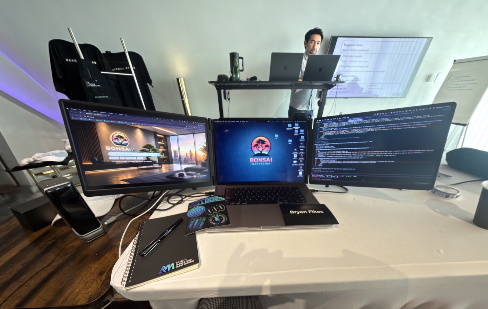 A multi-monitor workstation set up in a Las Vegas room, Bonsai branding on the displays and a laptop on the desk, with someone working at a standing desk behind it.